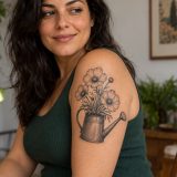 Regador preto e cinza com flores tatuado no braço de mulher adulta